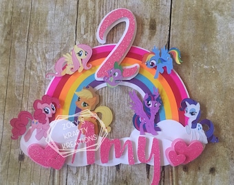 May include: A pink glittery number two cake topper with a rainbow and My Little Pony characters. The rainbow has a pink, orange, yellow, green, blue, and purple stripe. The characters include Pinkie Pie, Twilight Sparkle, Rainbow Dash, Fluttershy, Applejack, and Spike. The name "Amy" is written in pink glitter below the rainbow.