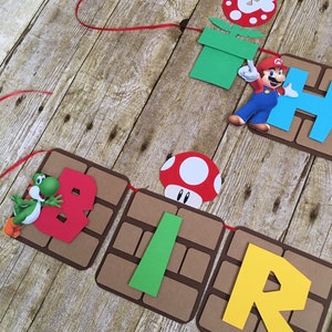 May include: A birthday banner with a Mario theme. The banner features a red, green and yellow colour scheme. The letters spell out "BIRTHDAY" and there are images of Mario, Yoshi and a mushroom.