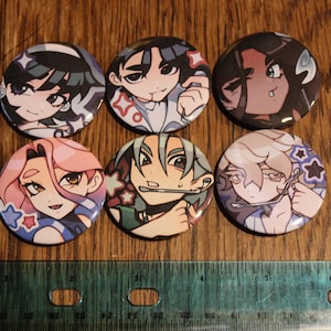 May include: Six round buttons with anime-style character illustrations. The buttons feature various characters with different hair colors and facial expressions. The buttons are approximately 1.5 inches in diameter and are set against a wooden background.