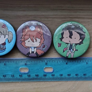 May include: Three circular pin-back buttons featuring anime-style character illustrations. The buttons are blue, purple, and green, each measuring approximately 1.25 inches in diameter as shown by the blue plastic ruler placed underneath them.