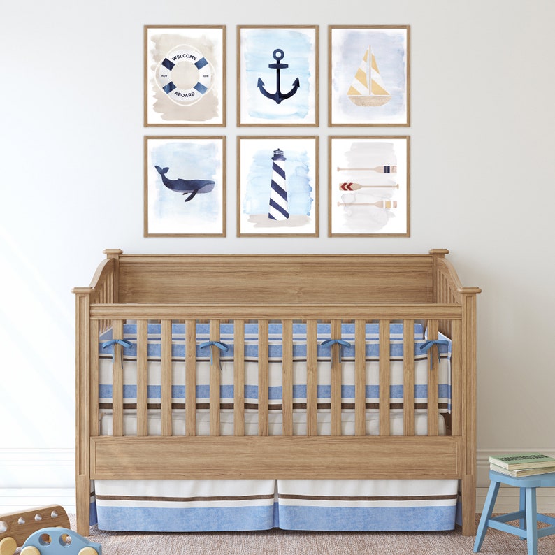May include: A wooden crib with a blue and white striped mattress and a blue and white striped skirt. The crib is decorated with six framed prints featuring nautical themes, including an anchor, a sailboat, a whale, a lighthouse, oars, and a life preserver with the text "Welcome Aboard".