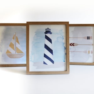 May include: Three framed watercolor paintings with nautical themes. The first painting features a sailboat with yellow and white stripes. The second painting features a lighthouse with blue and white stripes. The third painting features three oars with red, white, and blue accents.