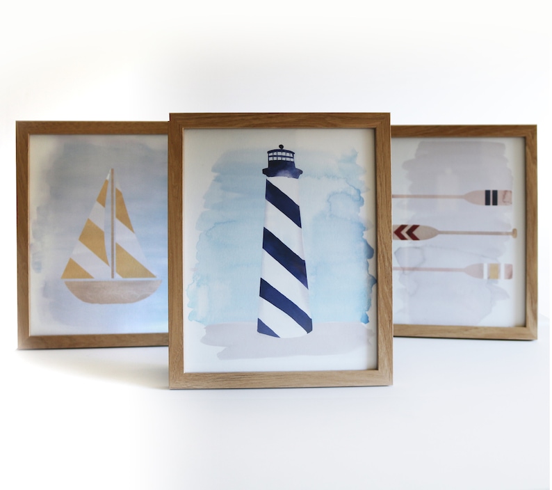 May include: Three framed watercolor paintings with nautical themes. The first painting features a sailboat with yellow and white stripes. The second painting features a lighthouse with blue and white stripes. The third painting features three oars with red, white, and blue accents.