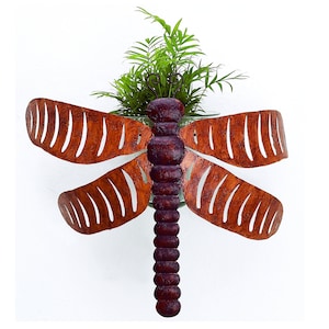 May include: A metal dragonfly wall planter with a rusty brown finish. The dragonfly has a green plant growing inside its body.