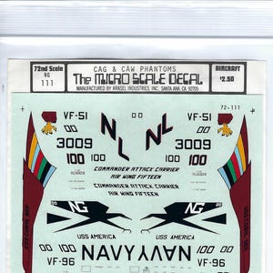 May include: A decal sheet for model aircraft featuring a variety of markings and insignia, including the words "Navy", "VF-96", "VF-114", and "NH". The sheet also includes the text "Commander Attack Carrier Air Wing Fifteen" and "USS America".