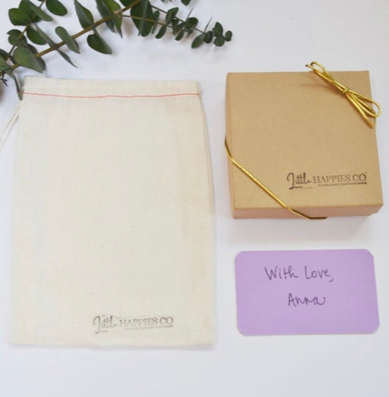 Peut inclure: Un petit sac en mousseline blanche avec un cordon rouge et le texte "Little Happies Co." imprim&eacute; au bas. Une bo&icirc;te brune avec un ruban dor&eacute; et le texte "Little Happies Co." imprim&eacute; sur le dessus. Une carte violette avec le texte manuscrit "Avec amour, Anna".