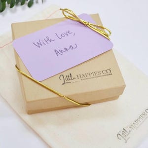K&ouml;nnte beinhalten: Eine kleine, quadratische, braune Geschenkbox mit einem hellvioletten Anh&auml;nger mit der Aufschrift "With Love, Anna". Die Box ist mit goldenem Garn gebunden und liegt auf einem cremefarbenen Stoffbeutel. Auf der Box steht "Little Happies Co".