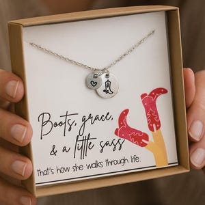 May include: A silver necklace with two charms, one heart-shaped and one with a cowboy boot design, presented in a gift box. The box features the text "Boots, grace, & a little sass" and a graphic of red cowboy boots.