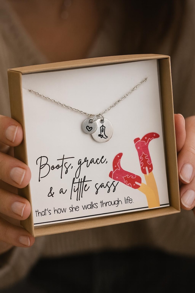 May include: A silver necklace with two charms, one heart-shaped and one with a cowboy boot design, presented in a gift box. The box features the text "Boots, grace, & a little sass" and a graphic of red cowboy boots.
