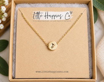 May include: A gold-toned necklace featuring a small circular pendant with a stamped musical note symbol. The necklace is displayed on a tan card inside a brown box. The card includes the text Little Happies Co and the website address.