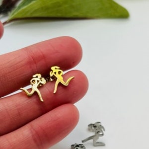 May include: Gold-colored and silver-colored running figure stud earrings. The earrings are in the shape of a person running. The earrings are small and delicate, suitable for everyday wear. The background is white with a green leaf.