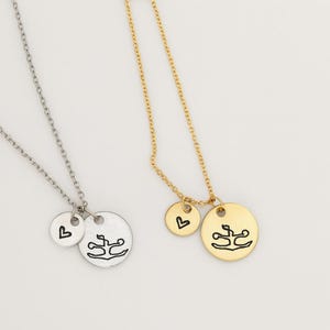 May include: Two necklaces: one silver, one gold. Each features a chain with a small round pendant and a larger circular pendant. The smaller pendant has a heart, and the larger pendant has a stylized design. Jewelry.