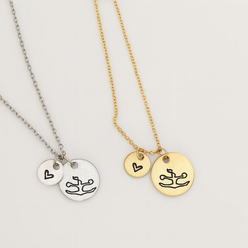 May include: Two necklaces: one silver, one gold. Each features a chain with a small round pendant and a larger circular pendant. The smaller pendant has a heart, and the larger pendant has a stylized design. Jewelry.
