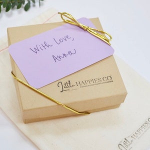 K&ouml;nnte beinhalten: Eine kleine braune Geschenkbox mit einer lila Karte, die mit einem goldenen Band gebunden ist. Die Karte lautet "Mit Liebe, Anna". Die Box tr&auml;gt das Logo "Little Happies Co."