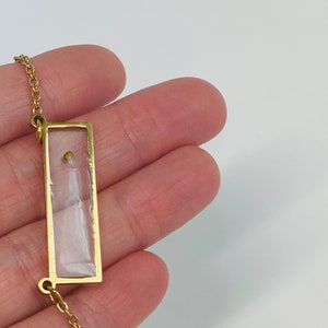 Pu&ograve; includere: Una collana con catena dorata e un ciondolo rettangolare. Il ciondolo &egrave; trasparente e contiene un piccolo perno dorato all'interno.