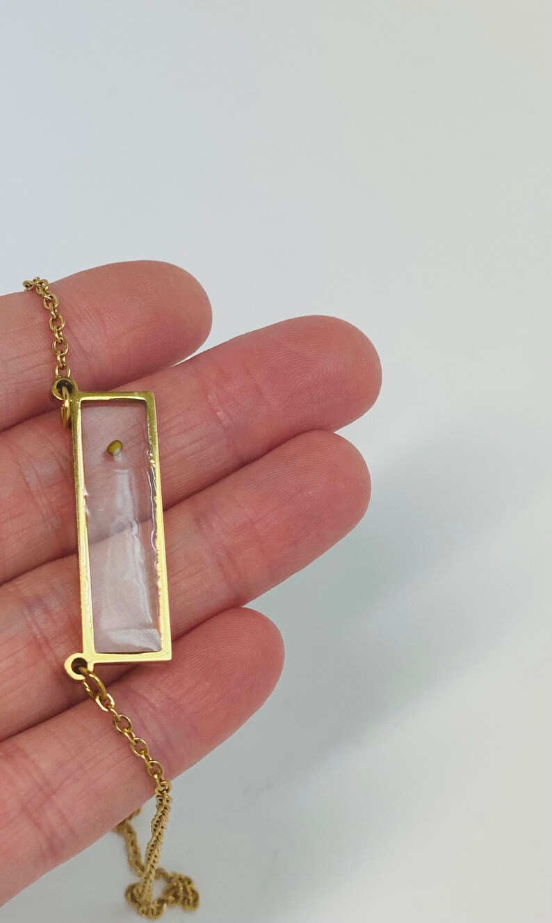 Pu&ograve; includere: Una collana con catena dorata e un ciondolo rettangolare. Il ciondolo &egrave; trasparente e contiene un piccolo perno dorato all'interno.