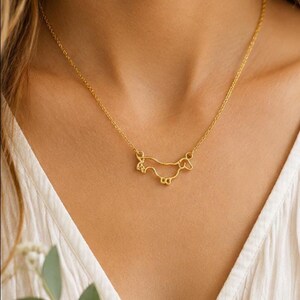 May include: A gold-toned necklace featuring a delicate wire-outline pendant shaped like a dachshund dog. The pendant is suspended from a thin, matching gold-toned chain that rests against the collarbone, showcasing a minimalist silhouette.