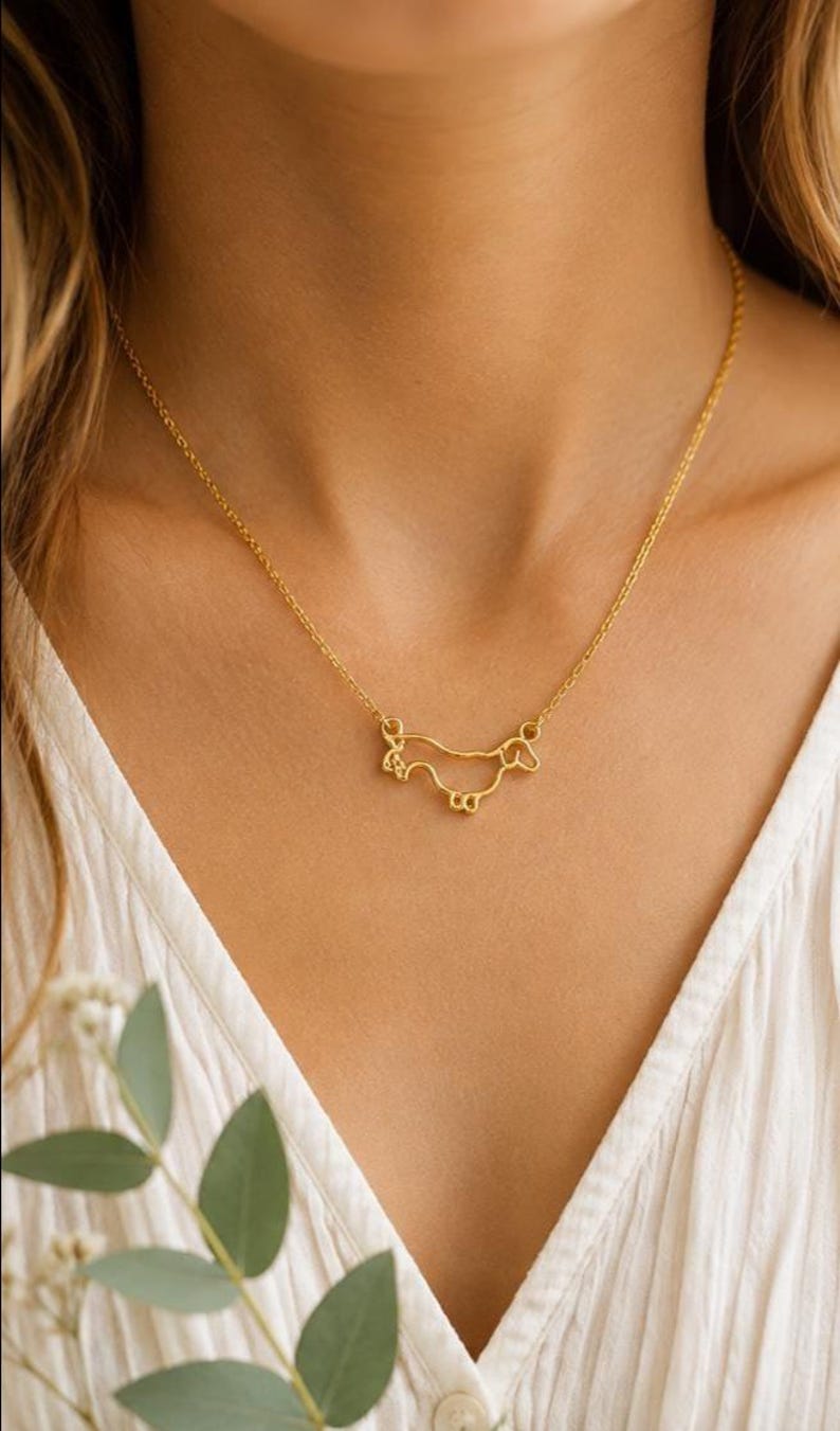 May include: A gold-toned necklace featuring a delicate wire-outline pendant shaped like a dachshund dog. The pendant is suspended from a thin, matching gold-toned chain that rests against the collarbone, showcasing a minimalist silhouette.