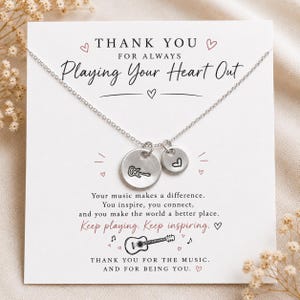 Könnte beinhalten: Eine silberfarbene Halskette mit zwei runden Anhängern, einer mit Gitarrengravur und einer mit einem Herz. Die Kette ist auf einer weißen Karte befestigt mit dem Text: THANK YOU FOR ALWAYS Playing Your Heart Out. Your music makes a difference.