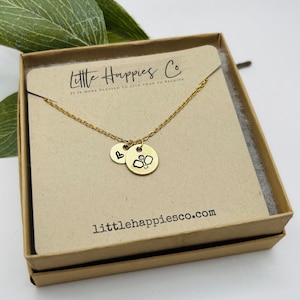 May include: Gold necklace with a heart charm and a charm featuring a pickleball design. The necklace is presented in a small brown box with the text "Little Happies Co. It is more blessed to give than to receive" and the website "littlehappiesco.com".
