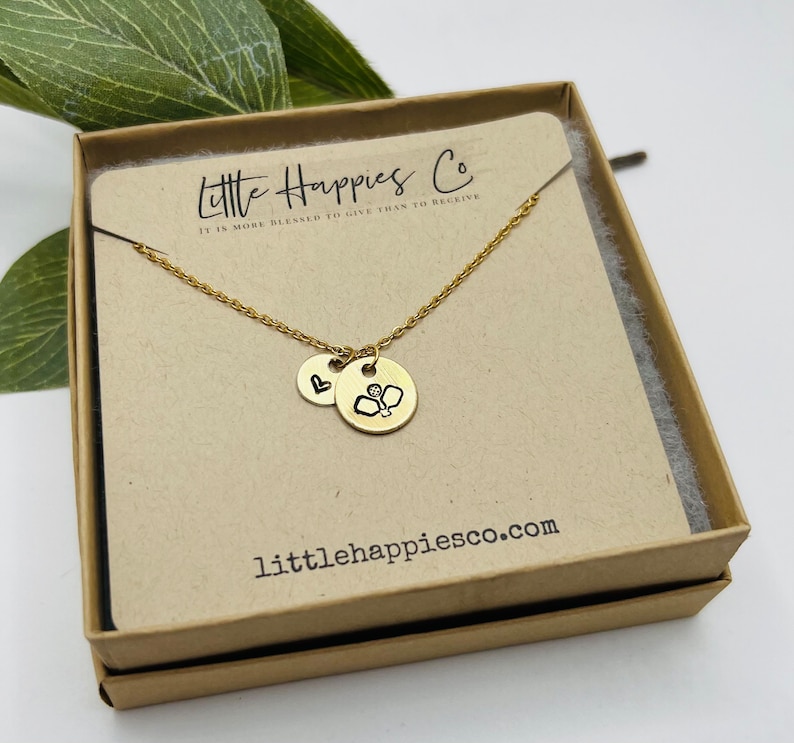 May include: Gold necklace with a heart charm and a charm featuring a pickleball design. The necklace is presented in a small brown box with the text "Little Happies Co. It is more blessed to give than to receive" and the website "littlehappiesco.com".