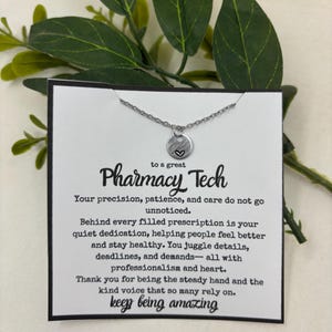 May include: A silver necklace with a small, round pendant featuring a heart design. The necklace is displayed on a white card with the text "to a great Pharmacy Tech" and a message of appreciation. Green leaves are in the background.