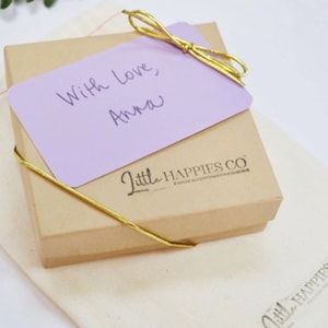 May include: A small, brown gift box with a purple card on top. The card has the handwritten message "With love, Anna". The box is tied with a gold ribbon and has the words "Little Happies Co." printed on the side.