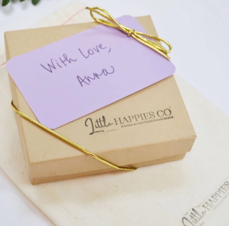 May include: A small, brown gift box with a purple card on top. The card has the handwritten message "With love, Anna". The box is tied with a gold ribbon and has the words "Little Happies Co." printed on the side.