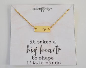 May include: Gold necklace with a small rectangular pendant that says "it takes a big heart to shape little minds". The pendant has a small heart cutout.