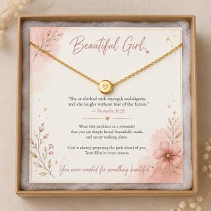 May include: A gold-colored necklace with a small circular pendant featuring a heart design, presented in a gift box. The box contains a card with the words "Beautiful Girl" and a quote from Proverbs 31:25. The card has a floral design.