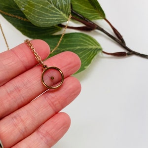 K&ouml;nnte beinhalten: Eine goldene Kettenkette mit einem kleinen runden Anh&auml;nger. Der Anh&auml;nger hat eine klare Glas- oder Plastikabdeckung und eine winzige goldene Perle im Inneren.