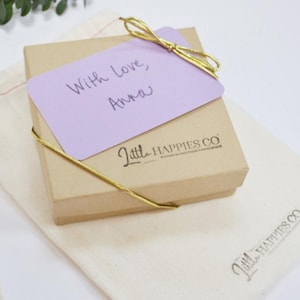 May include: A small, brown gift box with a purple card on top. The card has the handwritten message "With love, Anna". The box is tied with a gold ribbon and has the logo "Little Happies Co." printed on the side.