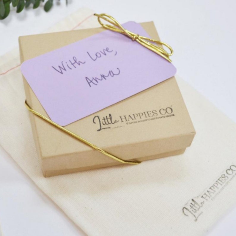 May include: A small, brown gift box with a purple card on top. The card has the handwritten message "With love, Anna". The box is tied with a gold ribbon and has the logo "Little Happies Co." printed on the side.