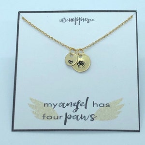 Puede incluir: Collar de oro con dos dijes, un coraz&oacute;n y una huella de pata. El collar est&aacute; sobre una tarjeta blanca con el texto "my angel has four paws".