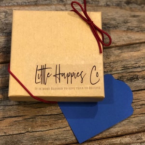 Puede incluir: Una peque&ntilde;a caja de cart&oacute;n marr&oacute;n con una cinta roja atada alrededor. La caja tiene una etiqueta blanca con el texto "Little Happies Co. It is more blessed to give than to receive." impreso en ella.
