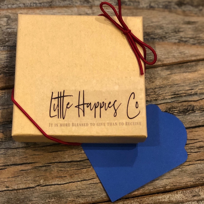 Puede incluir: Una peque&ntilde;a caja de cart&oacute;n marr&oacute;n con una cinta roja atada alrededor. La caja tiene una etiqueta blanca con el texto "Little Happies Co. It is more blessed to give than to receive." impreso en ella.