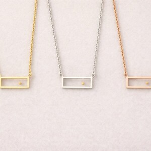 May include: Three necklaces featuring thin metal chains and rectangular pendants. Each pendant is an open frame containing a single small dot positioned off-center. The set includes one gold-tone, one silver-tone, and one rose gold-tone necklace.