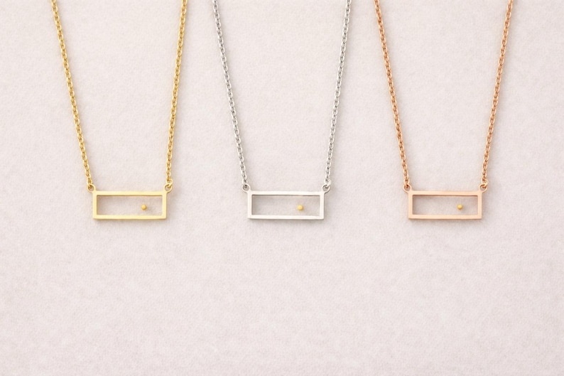 May include: Three necklaces featuring thin metal chains and rectangular pendants. Each pendant is an open frame containing a single small dot positioned off-center. The set includes one gold-tone, one silver-tone, and one rose gold-tone necklace.