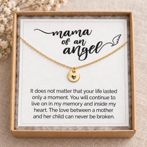 May include: A gold-toned necklace with a small circular pendant featuring a black heart engraving. The necklace is displayed on a white card inside a brown box, with the text "mama of an angel" and a memorial message printed on the card.