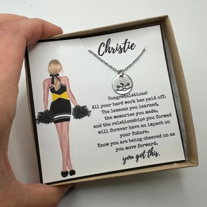 May include: A silver necklace with a round pendant featuring a cheerleader graphic, presented in a gift box. The box includes a card with a cheerleader illustration and the text "Christie" and "you got this."