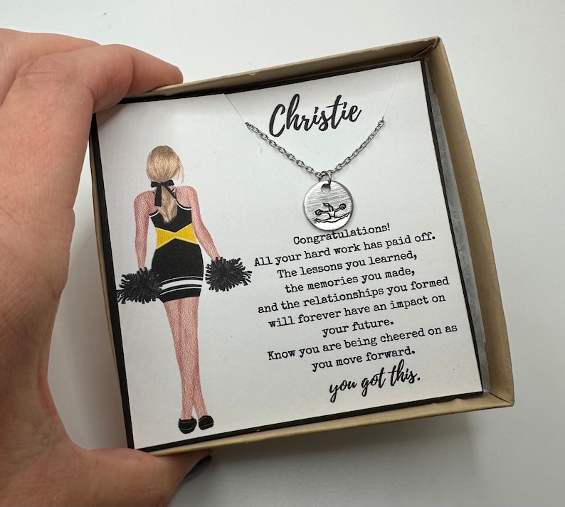May include: A silver necklace with a round pendant featuring a cheerleader graphic, presented in a gift box. The box includes a card with a cheerleader illustration and the text "Christie" and "you got this."