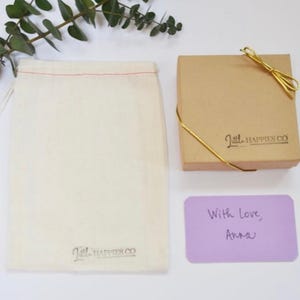 May include: An overhead shot shows a beige gift box tied with gold string, a drawstring bag, and a lavender card with handwritten text. The bag and box have the logo "Little Happies Co." printed on them. Eucalyptus sprigs are in the upper left.