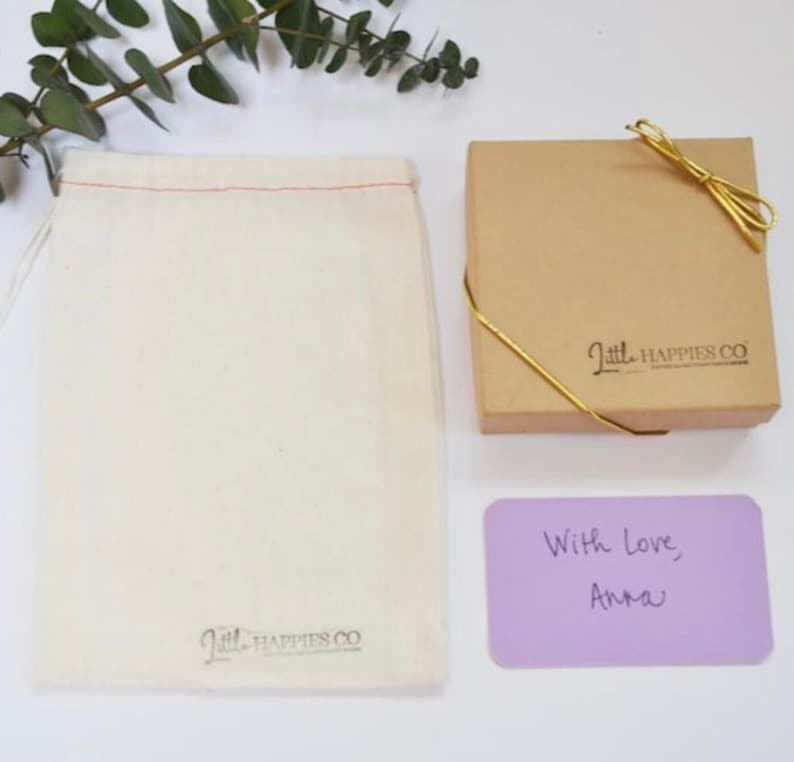 May include: An overhead shot shows a beige gift box tied with gold string, a drawstring bag, and a lavender card with handwritten text. The bag and box have the logo "Little Happies Co." printed on them. Eucalyptus sprigs are in the upper left.