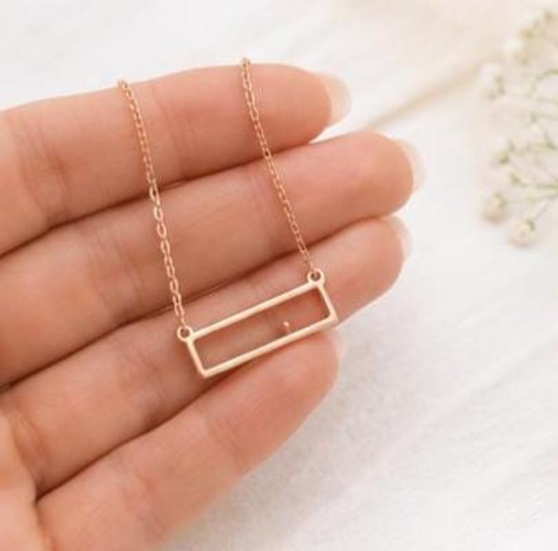 May include: A rose gold-toned necklace featuring a delicate chain and a rectangular pendant frame. The pendant is an open, thin-walled rectangle with a small, singular dot accent positioned inside the lower horizontal bar of the frame.