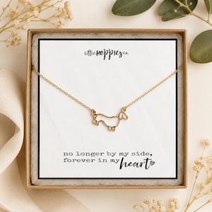 May include: A gold-toned necklace featuring a delicate outline pendant of a dachshund dog. The necklace is displayed on a white card inside a brown box, with the text "littlehappiesco." and "no longer by my side, forever in my heart".
