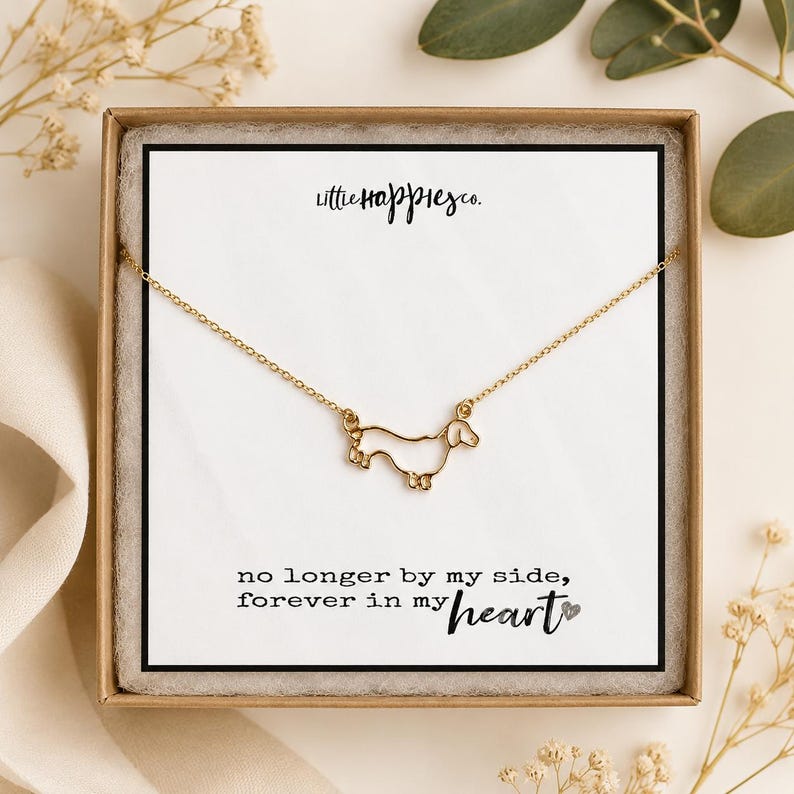 May include: A gold-toned necklace featuring a delicate outline pendant of a dachshund dog. The necklace is displayed on a white card inside a brown box, with the text "littlehappiesco." and "no longer by my side, forever in my heart".