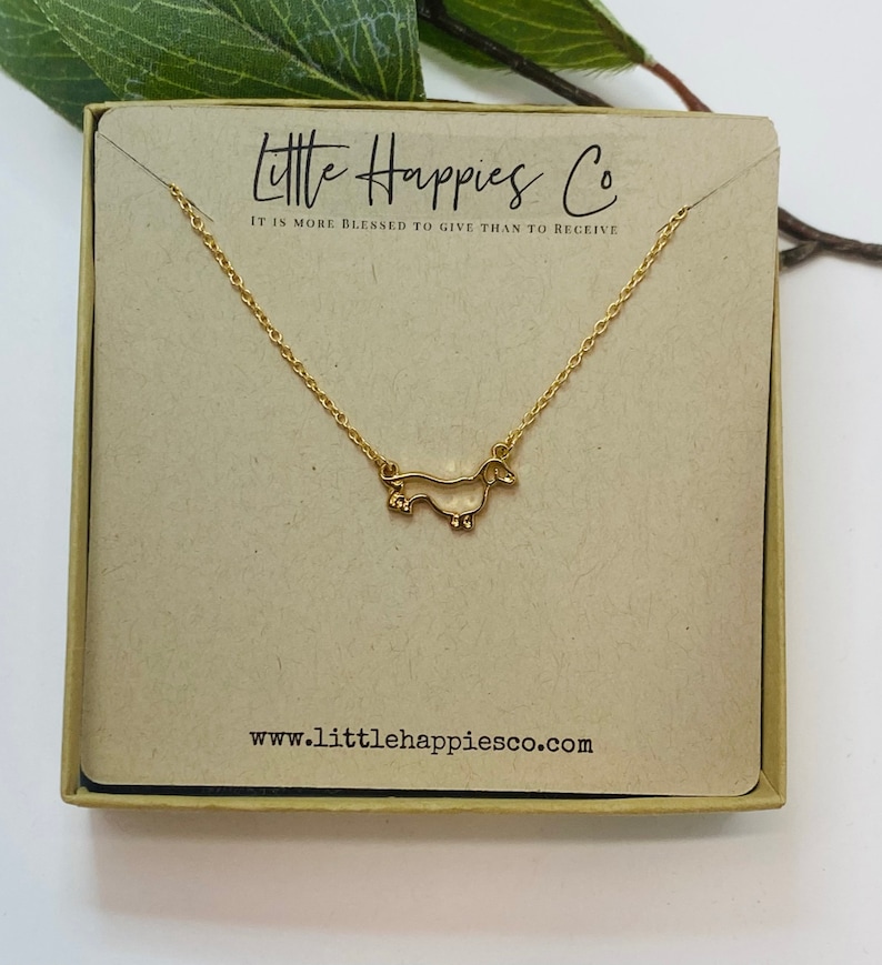 Peut inclure: Collier en or en forme de dachshund dans une petite bo&icirc;te en carton avec le texte "Little Happies Co. Il est plus b&eacute;ni de donner que de recevoir" et le site web "www.littlehappiesco.com".
