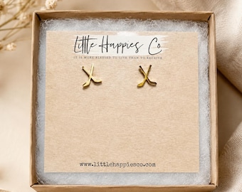 May include: A pair of gold-toned stud earrings shaped like crossed hockey sticks, displayed on a rectangular beige card inside a brown cardboard box. The card features the text Little Happies Co and the website www.littlehappiesco.com.