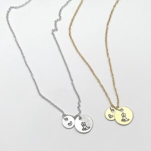 May include: Two delicate necklaces, one silver and one gold, each featuring a small charm with a heart and a round charm with a cowboy boot design. The necklaces are displayed on a white surface.