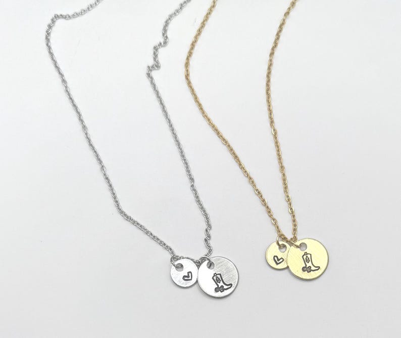 May include: Two delicate necklaces, one silver and one gold, each featuring a small charm with a heart and a round charm with a cowboy boot design. The necklaces are displayed on a white surface.